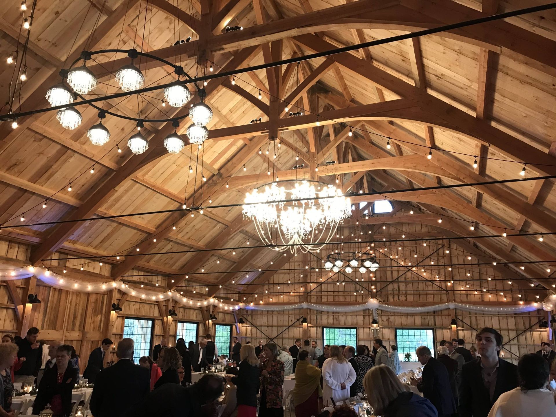 Rustic banquet hall with chandeliers, wooden beams, and guests mingling under a high vaulted ceiling