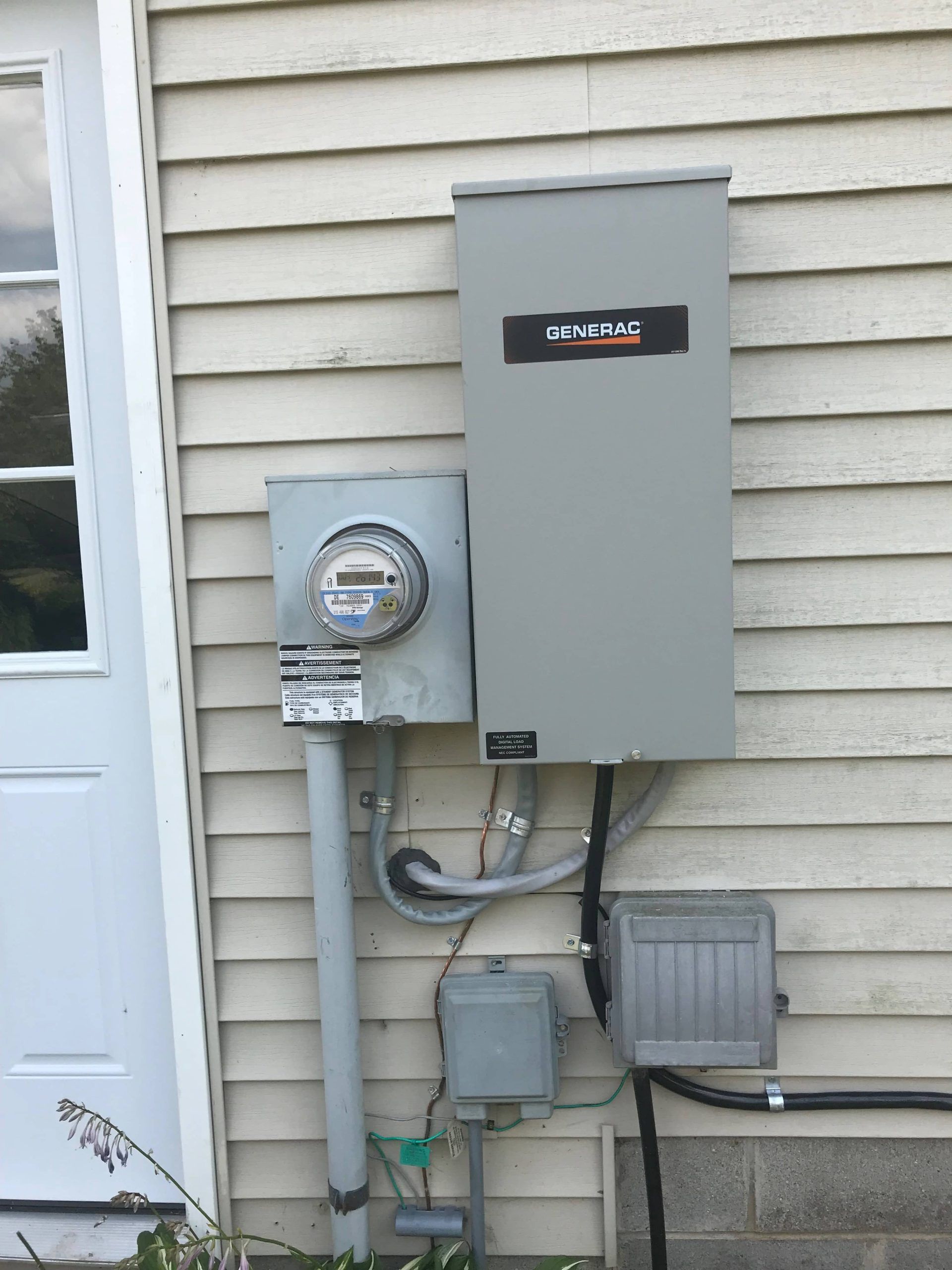 Outdoor electrical meter and gray utility boxes mounted on a house exterior.