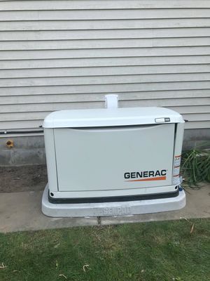White Generac backup generator on a concrete pad beside a house wall and grass.