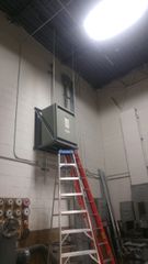 Industrial room with a tall ladder under a ceiling-mounted electrical unit and bright overhead light.