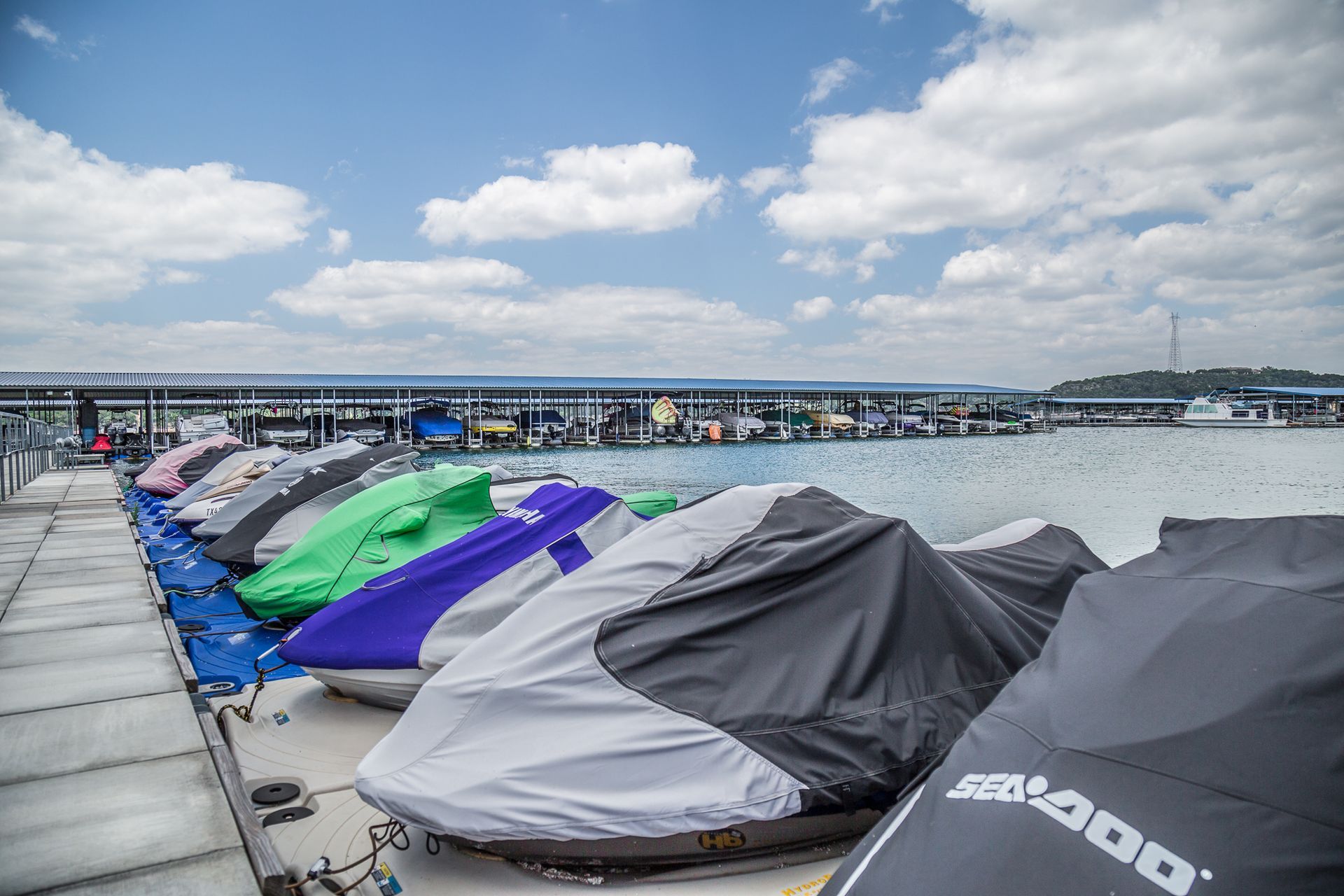 Jet skis covered with colorful tarps docked on a pier with a marina in the background.
