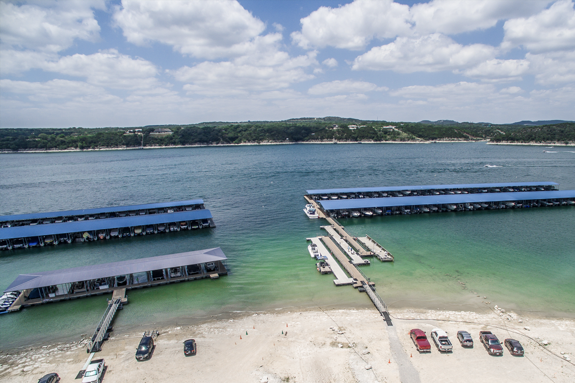 Aerial view of a lake with boat docks and a beach; blue water, blue roofs, cars parked on the shore, sunny day.