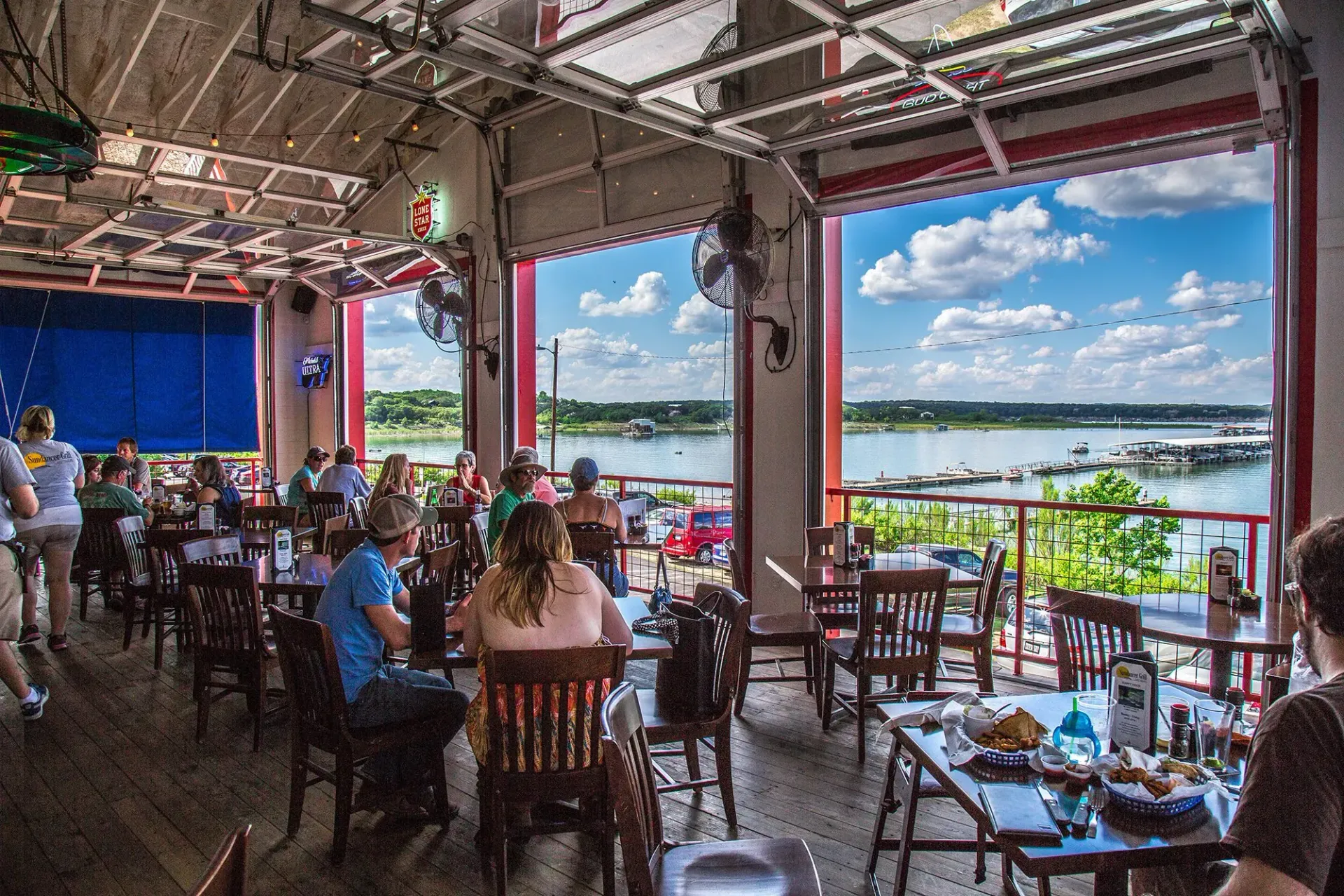 Restaurant patio with a lake view; people dine at tables indoors and outdoors.