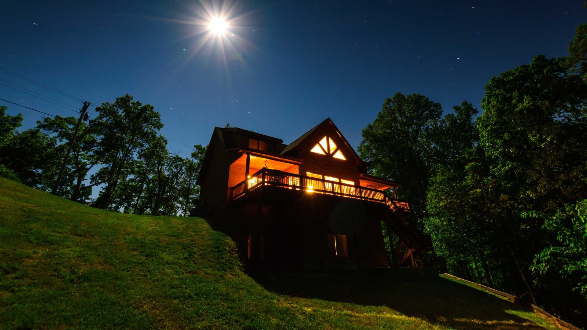 A cabin is lit up at night with a full moon in the sky.