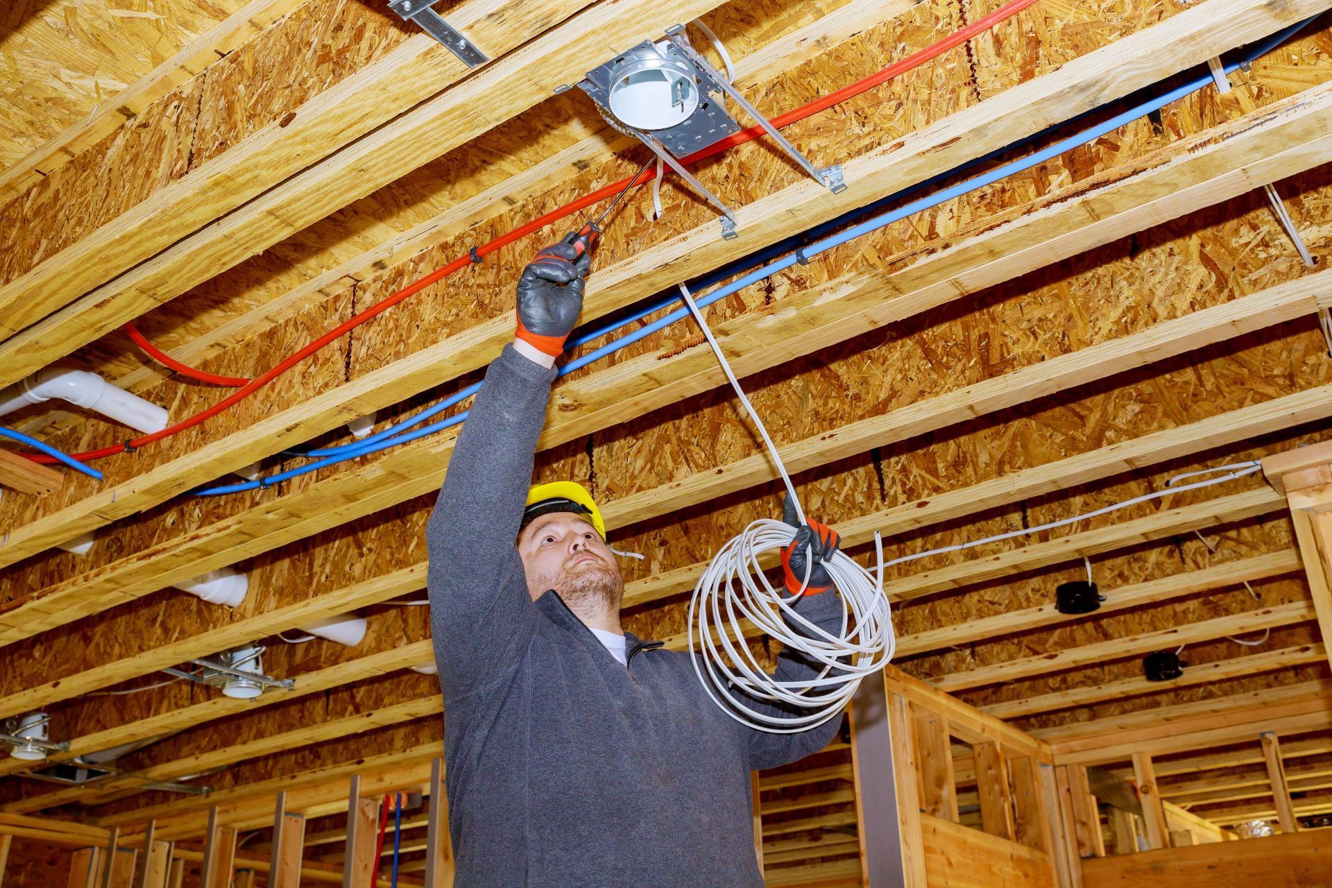 A construction worker in a hard hat installs electrical wiring for a ceiling light in an unfinished wood-frame building.