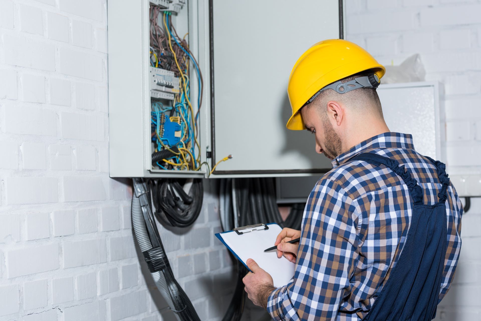 Electrician in yellow hard hat, inspecting electrical panel, writing on clipboard.