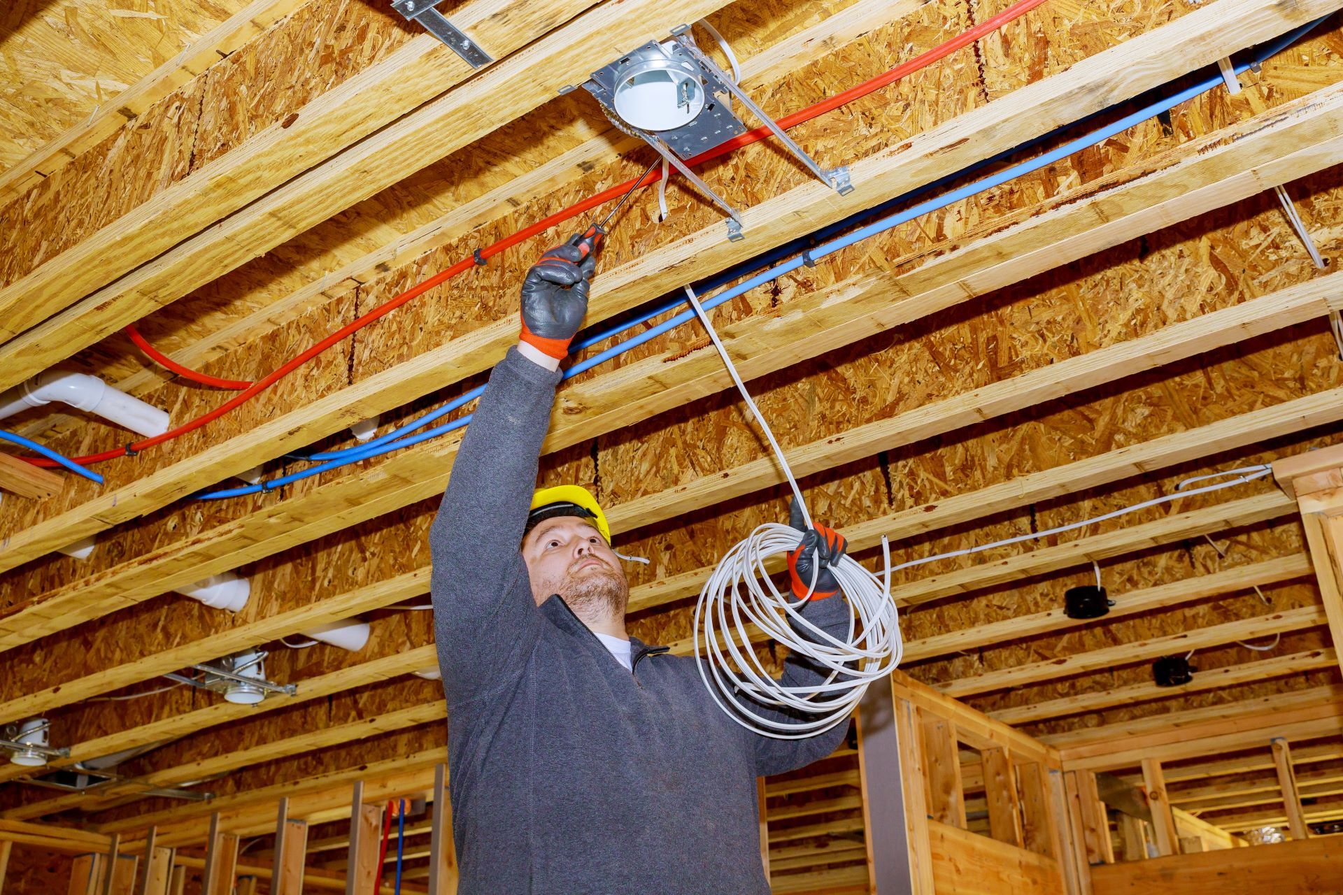 An electrician wearing a hard hat installs electrical wiring in the exposed wooden ceiling of a house under construction.