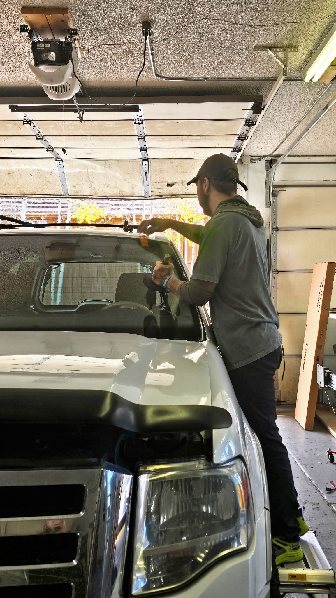 Person in a garage leaning over the hood of a white SUV, adjusting something on the windshield.