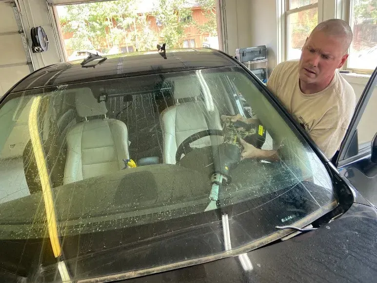 Man washing a black car windshield in a garage with soapy water and a hose