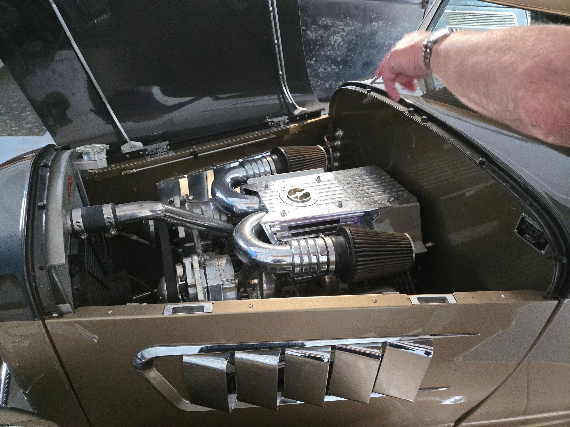 Open car hood revealing a custom engine with chrome and black components. A hand is reaching into frame.
