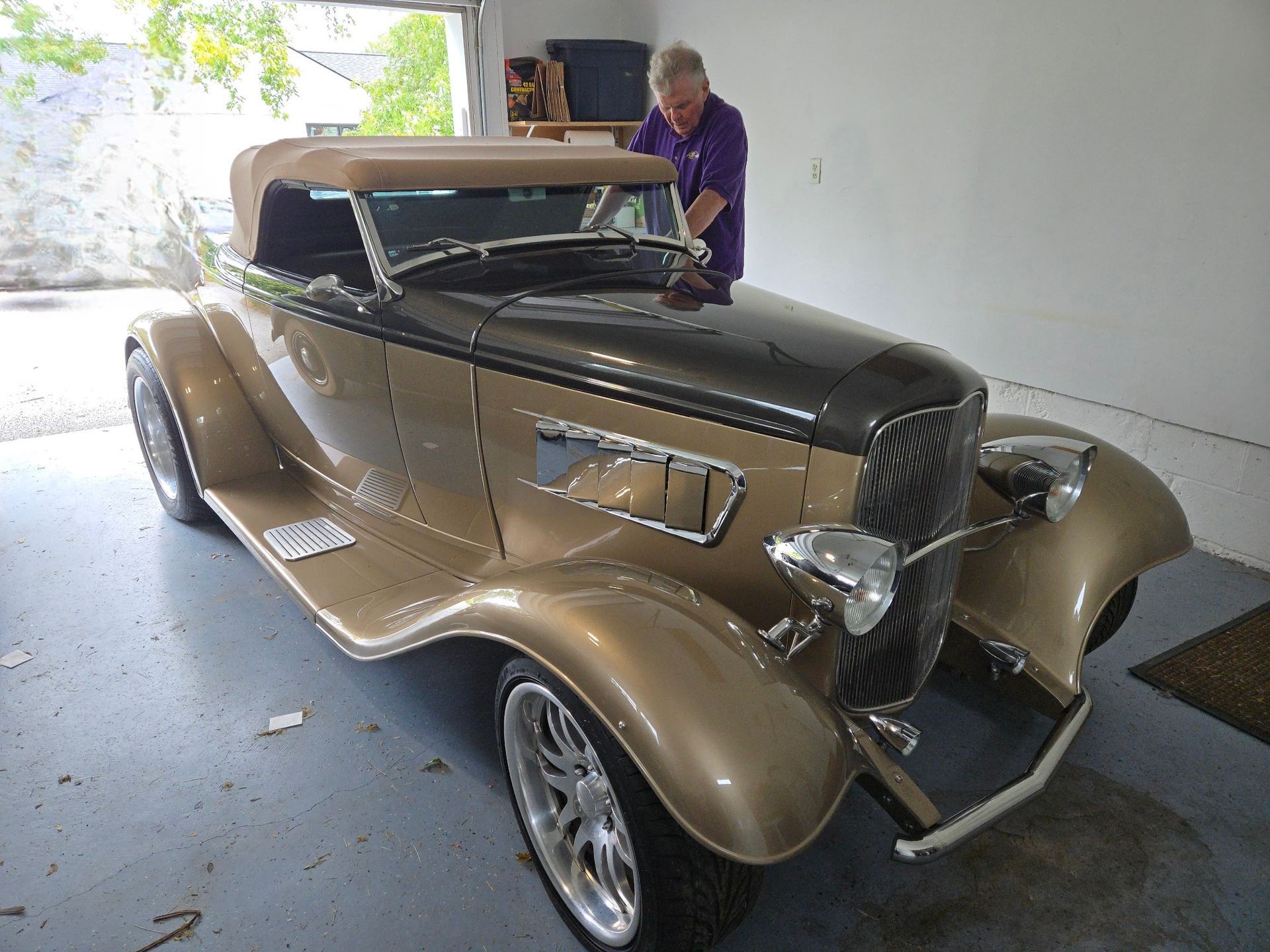 A gold and black vintage convertible car with a man in purple shirt beside it in a garage.