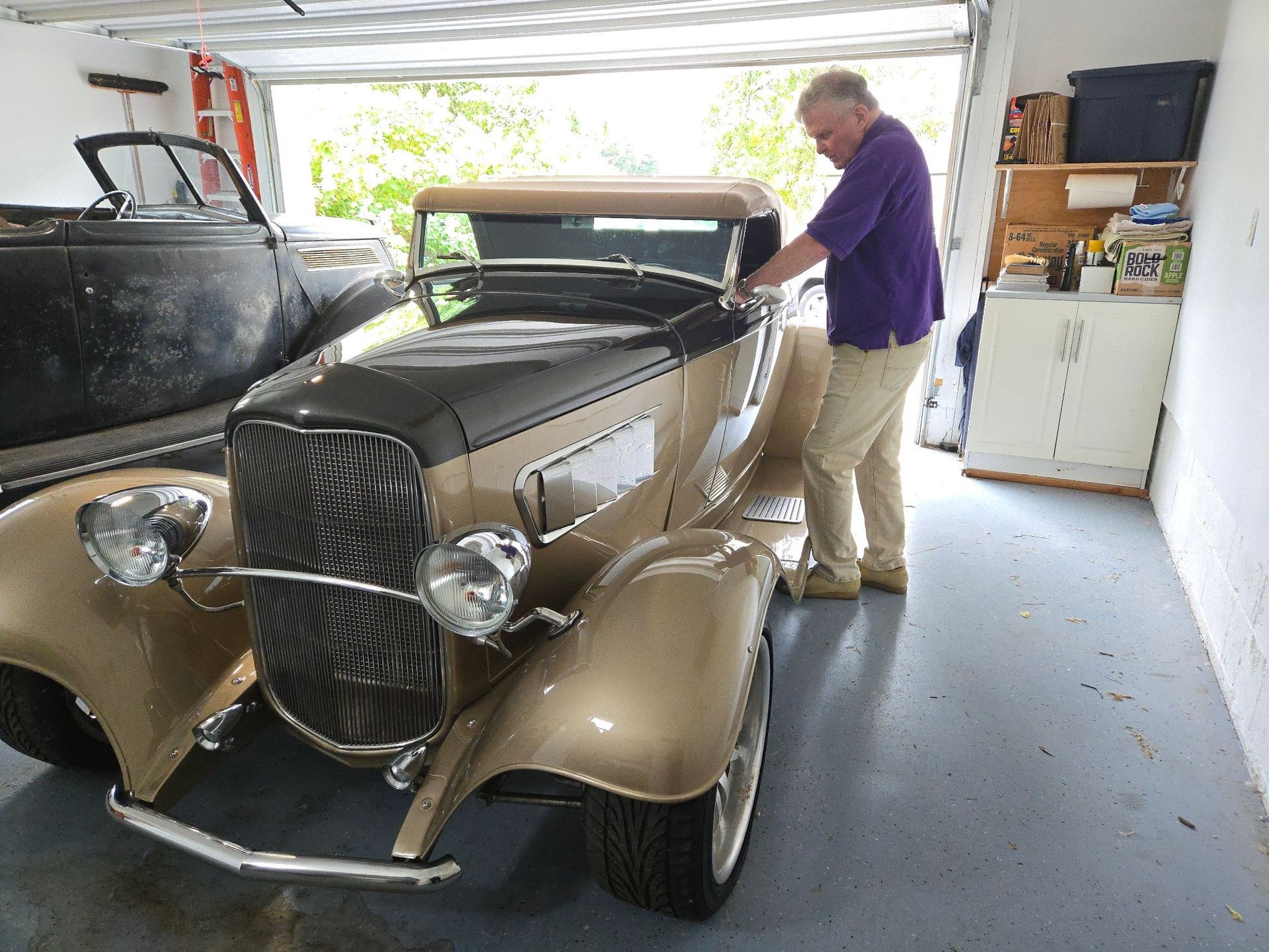 Man in purple shirt tending to a gold and black classic car in a garage.