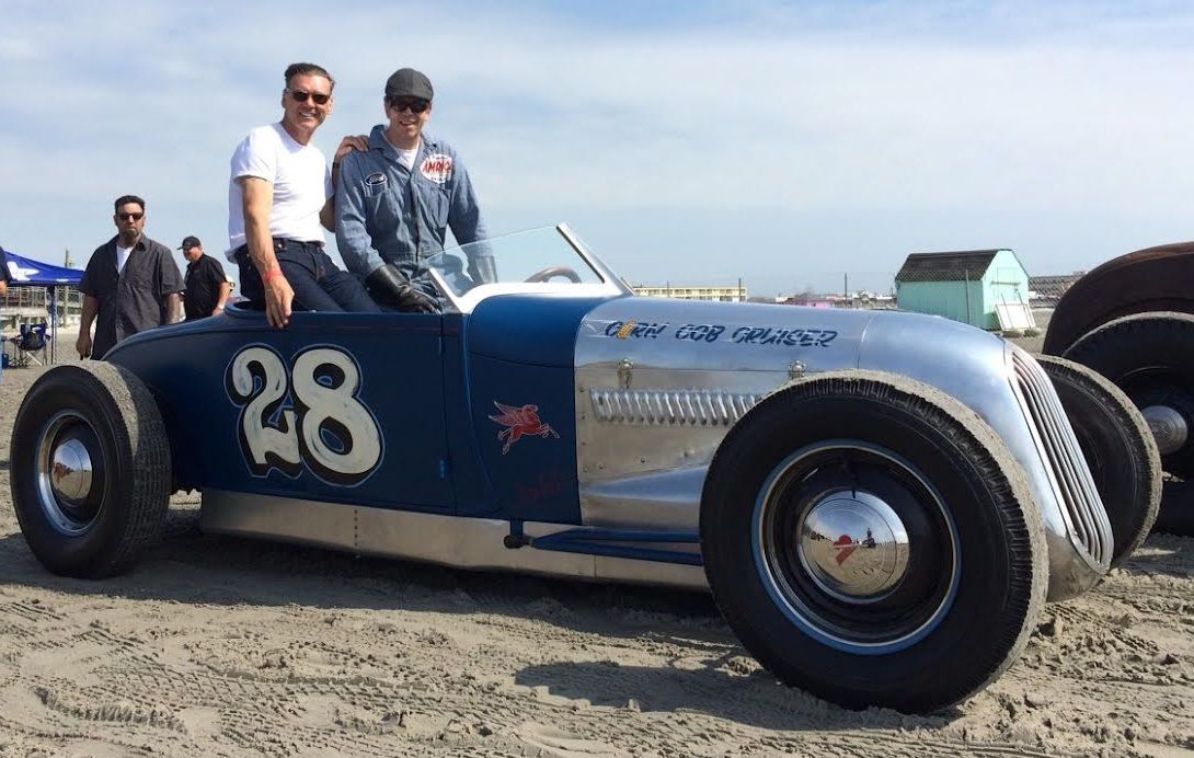 Two men pose beside a vintage blue and silver race car on a beach, number 