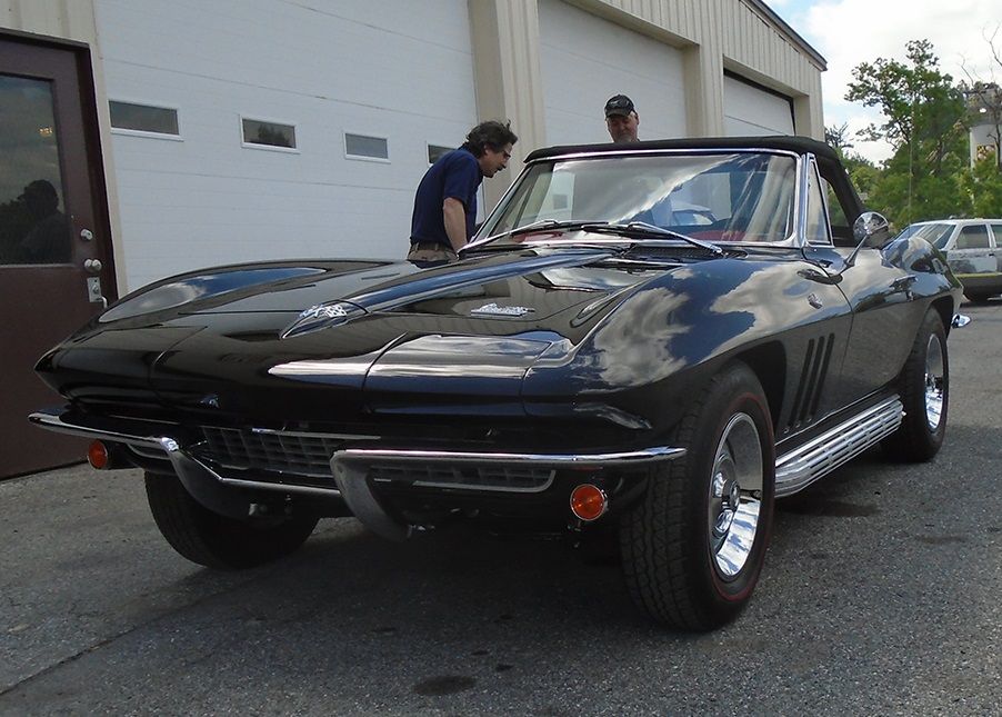 Black convertible Corvette parked near a garage; two people inspecting the car.