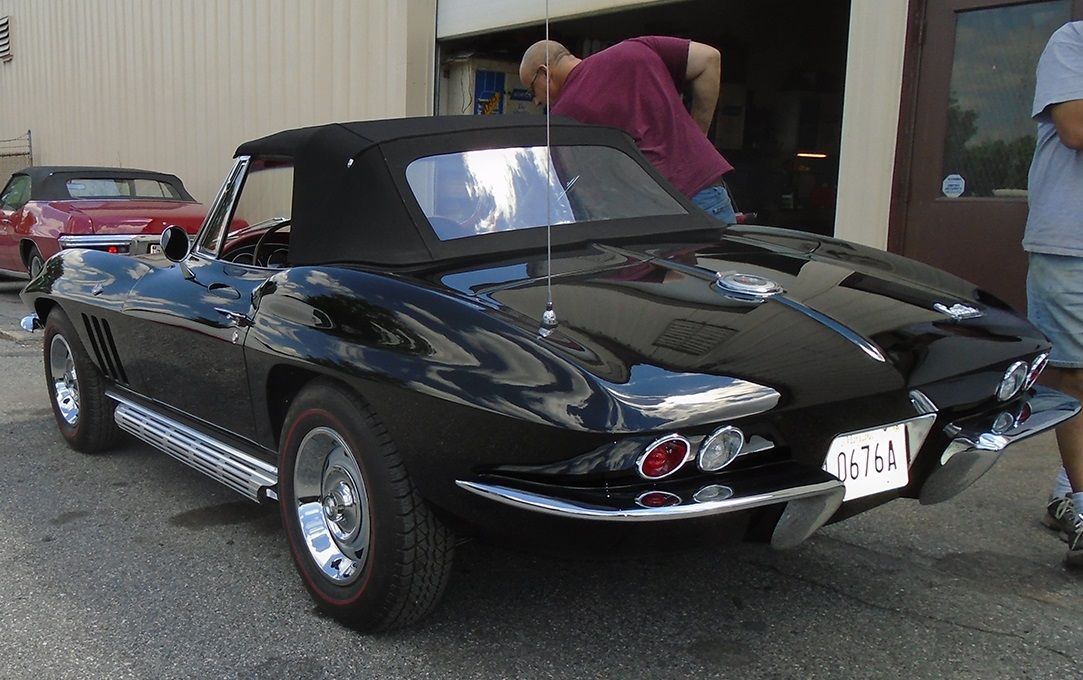 Black convertible Corvette with chrome accents, parked outdoors with people nearby.