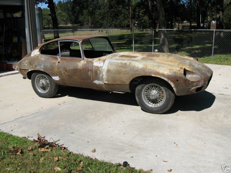 A rusty Jaguar E-Type coupe needing restoration, parked on a driveway.