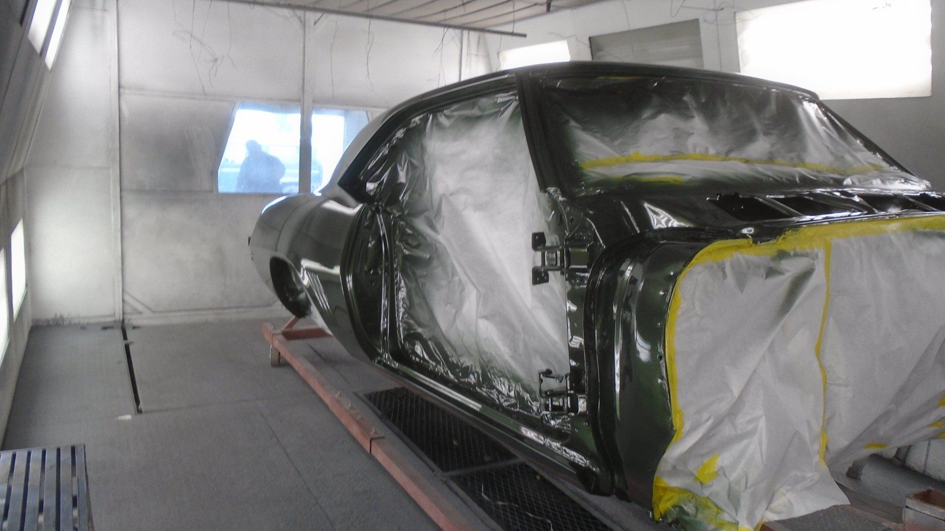 Classic car being painted green inside a spray booth. Door and other areas are masked off with white plastic.