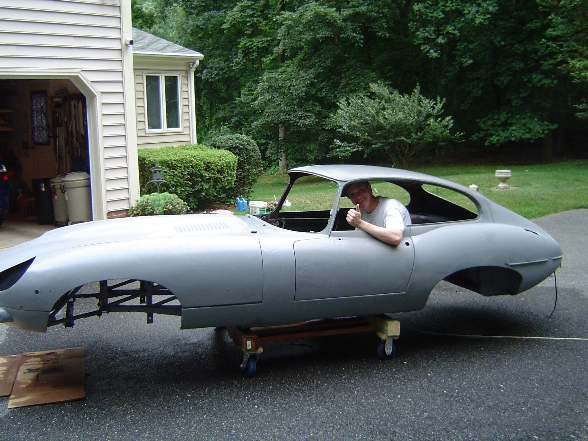 Man in gray car frame, body on a dolly. Garage and house in the background.
