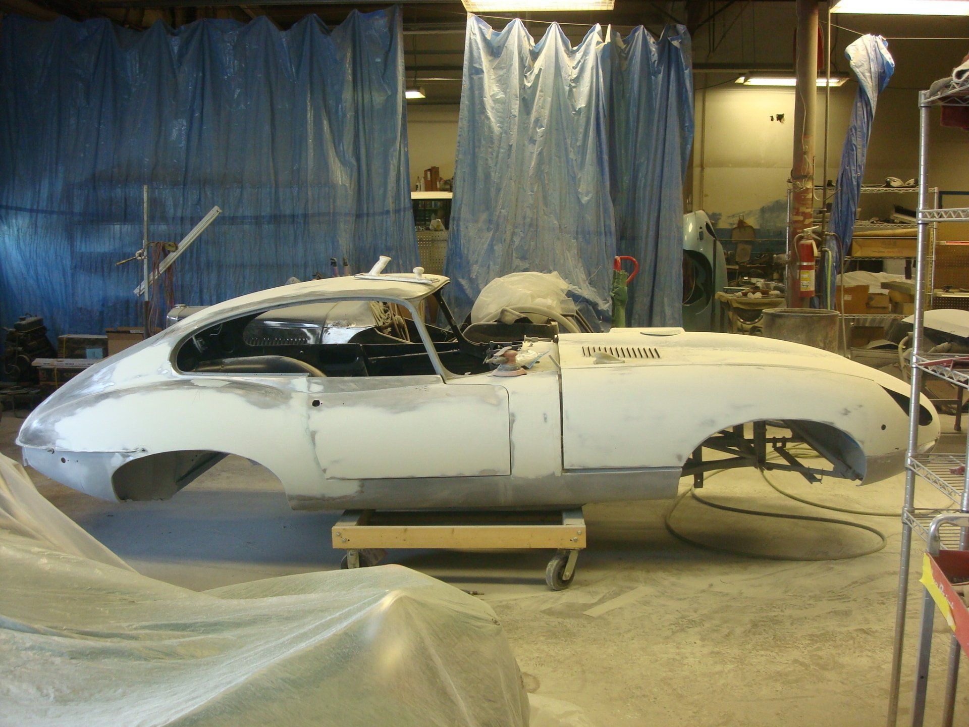 White Jaguar E-Type in a workshop, undergoing restoration. Body panels are primed, car is on a wooden stand.