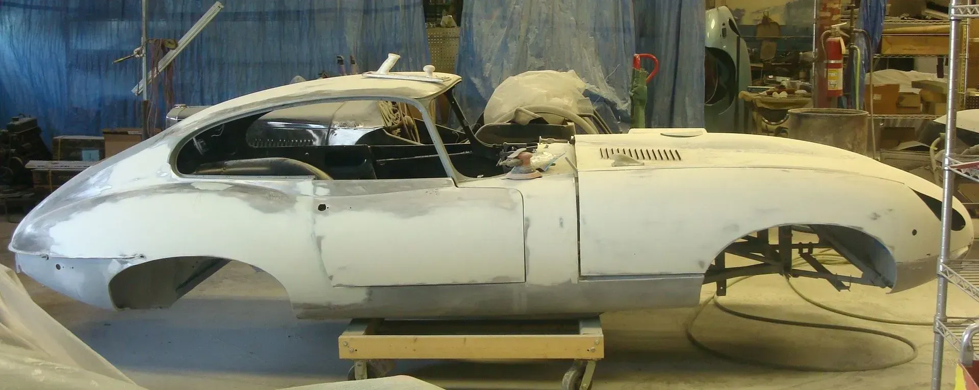 Jaguar E-Type undergoing restoration; white car body on wooden supports in a workshop.