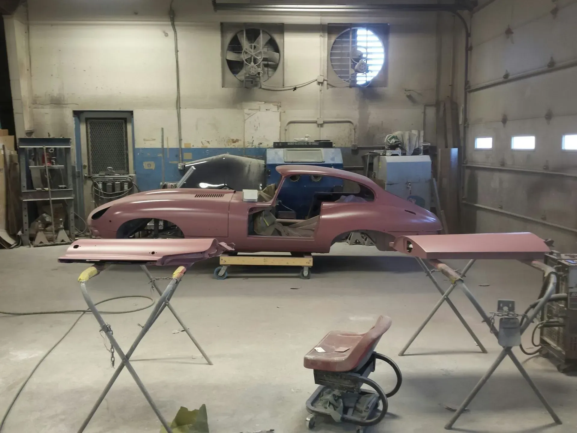 Classic car undergoing restoration in a workshop; pink body, detached panels, and tools visible.