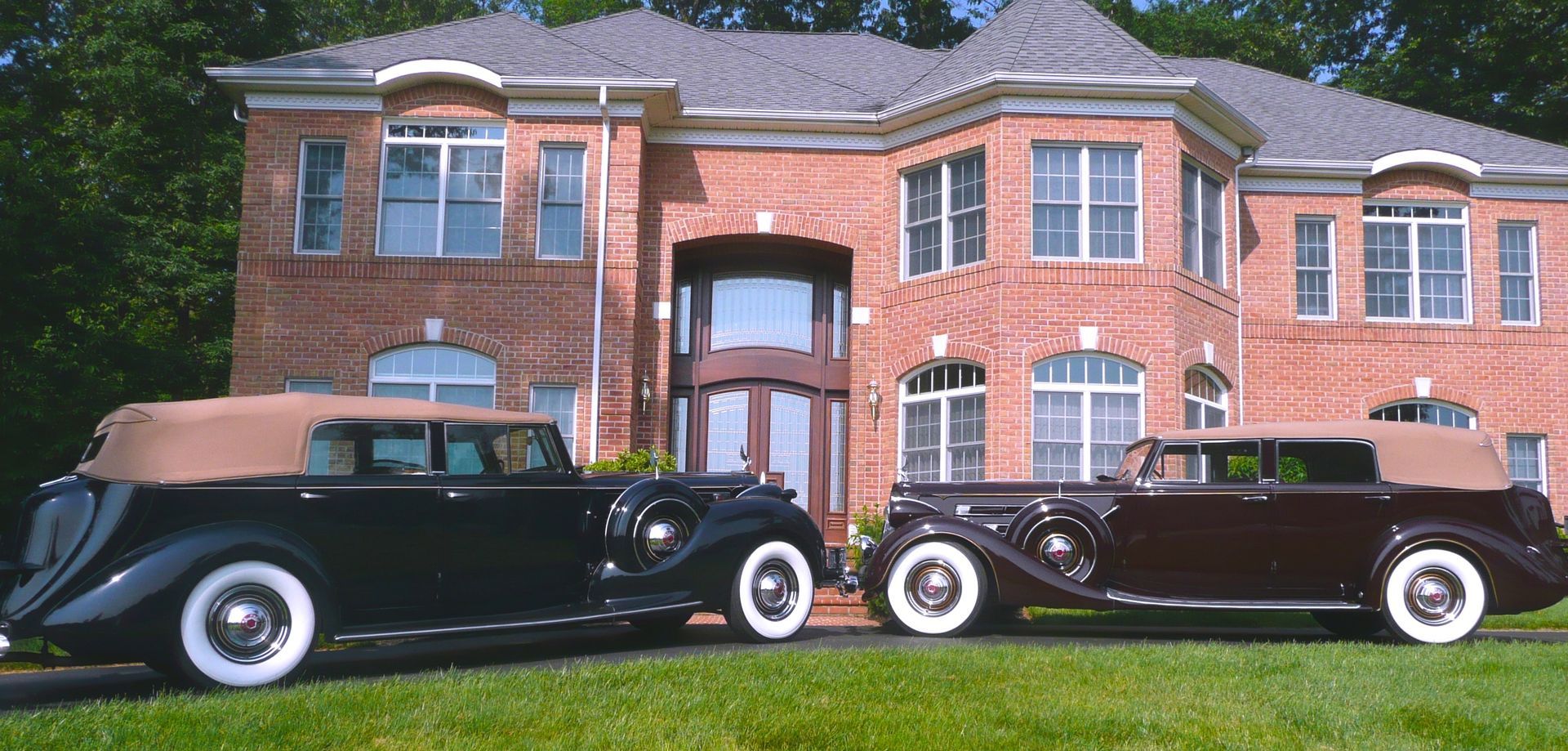Two vintage convertibles parked on a lawn in front of a brick house.