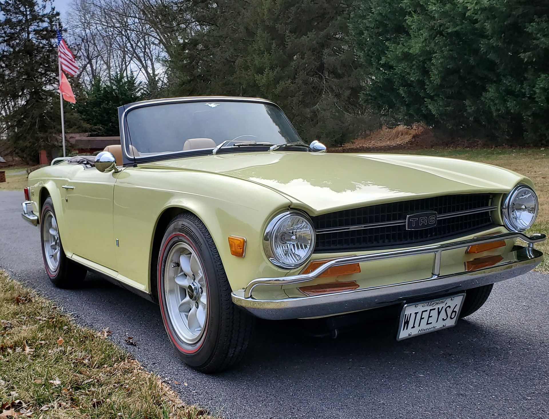 Yellow Triumph TR6 convertible parked on a driveway with an American flag in the background.