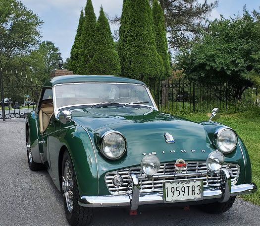Green vintage Triumph TR3 sports car parked on asphalt, with trees in background.