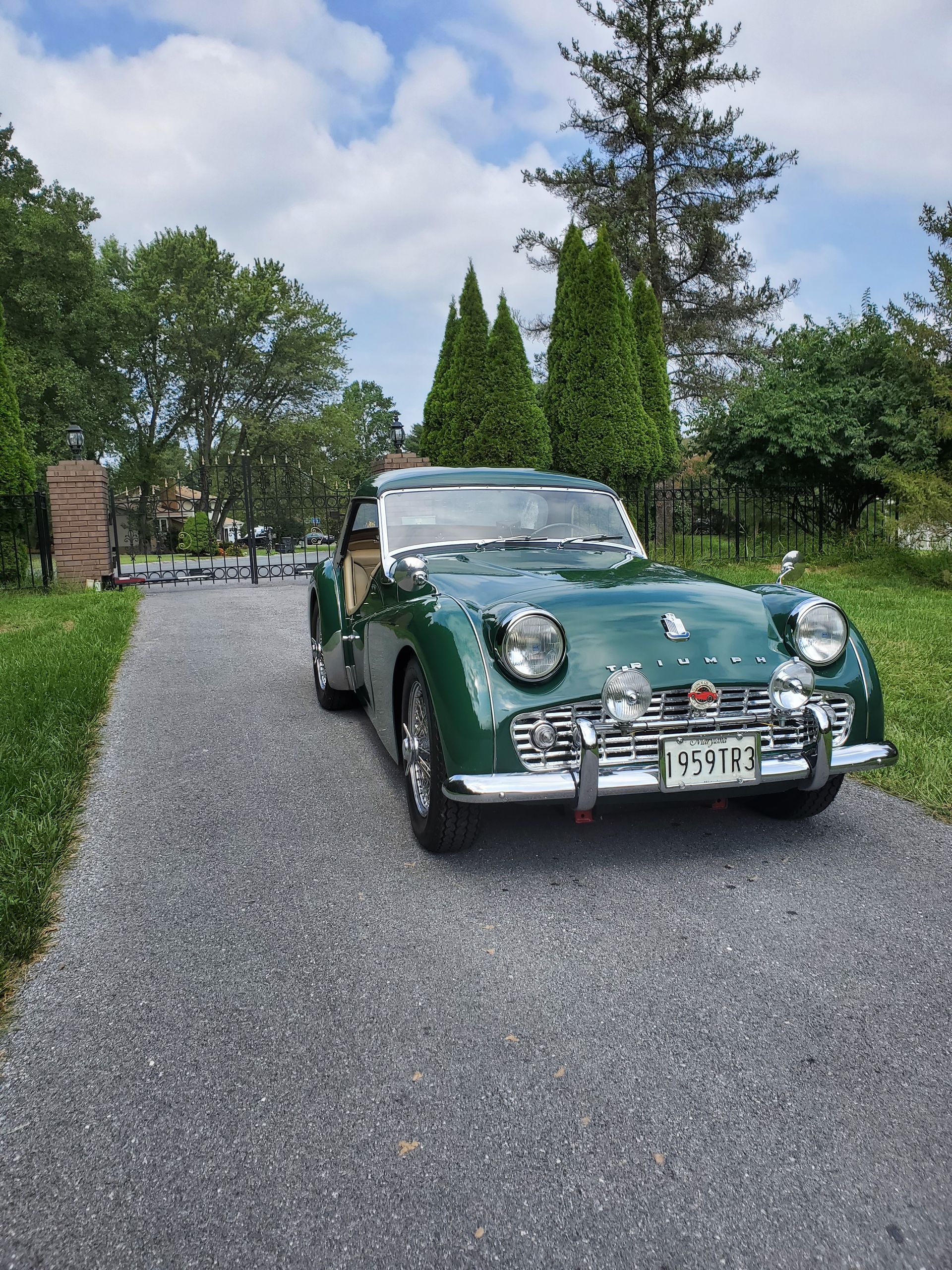 Green vintage Triumph sports car parked on a paved driveway, trees in background, sunny day.