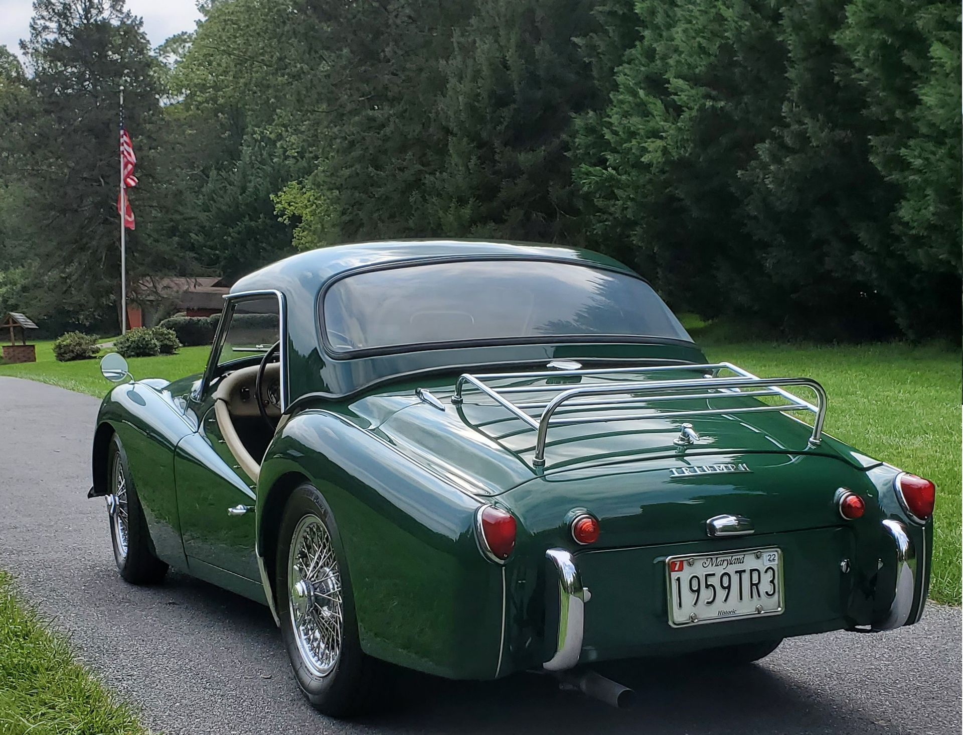 Green Triumph TR3 sports car with a hardtop, parked on a paved driveway near trees and a flagpole.