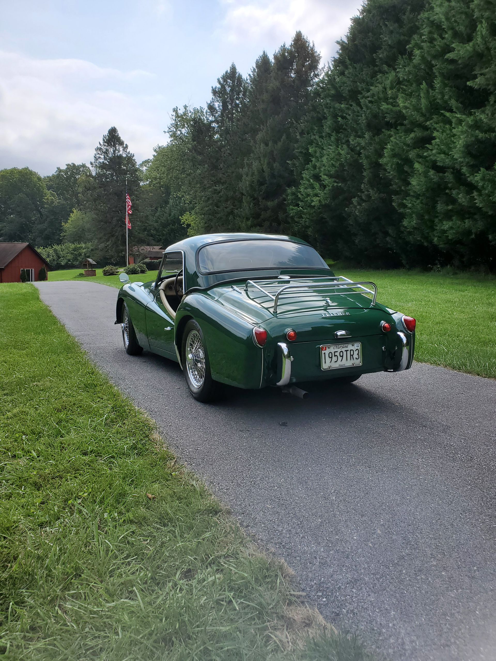 Green Triumph sports car on a paved driveway, trees in the background, a small building to the left.