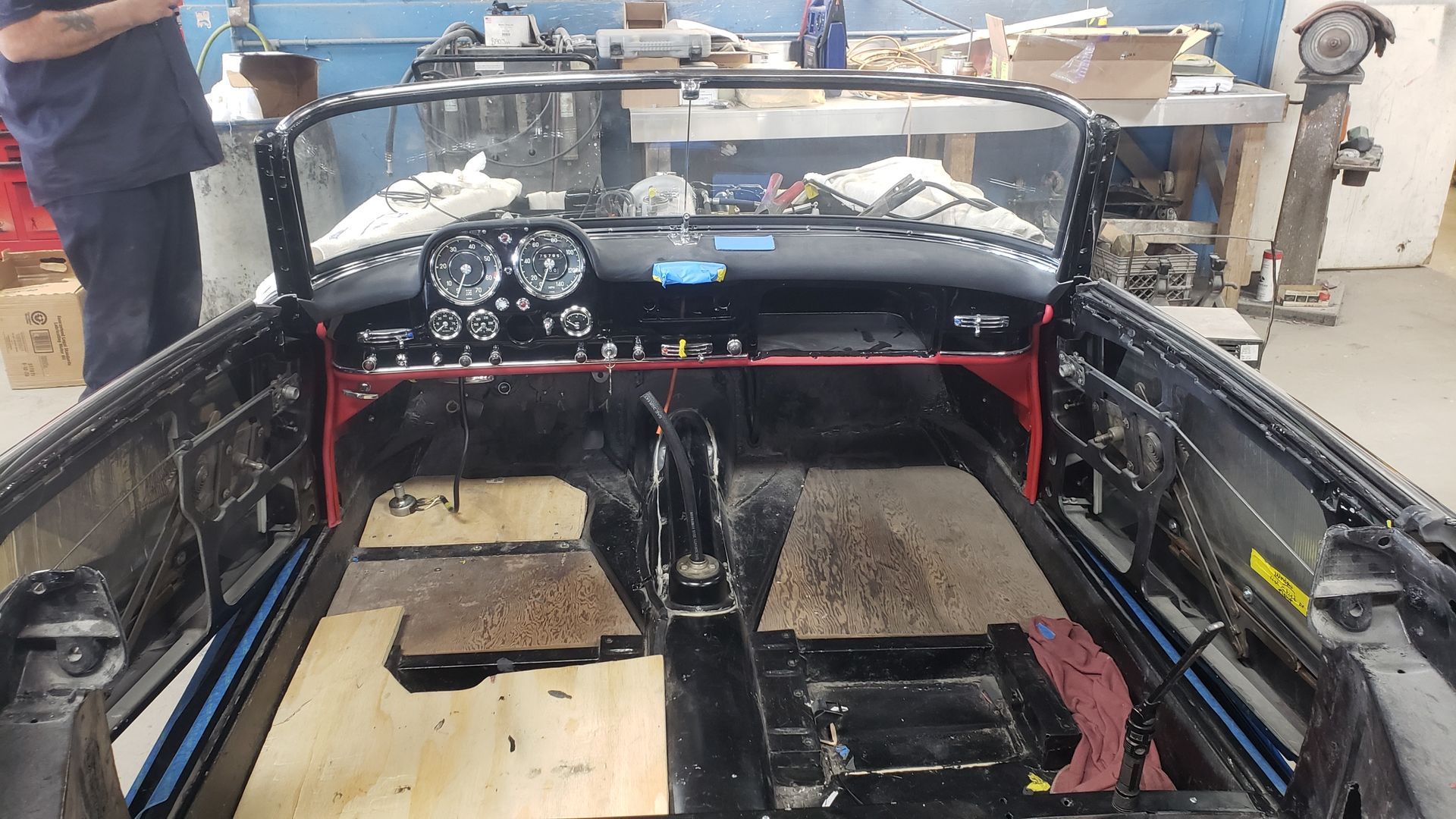 Interior of a vintage convertible car under restoration, black dashboard, red frame, wooden floorboards.