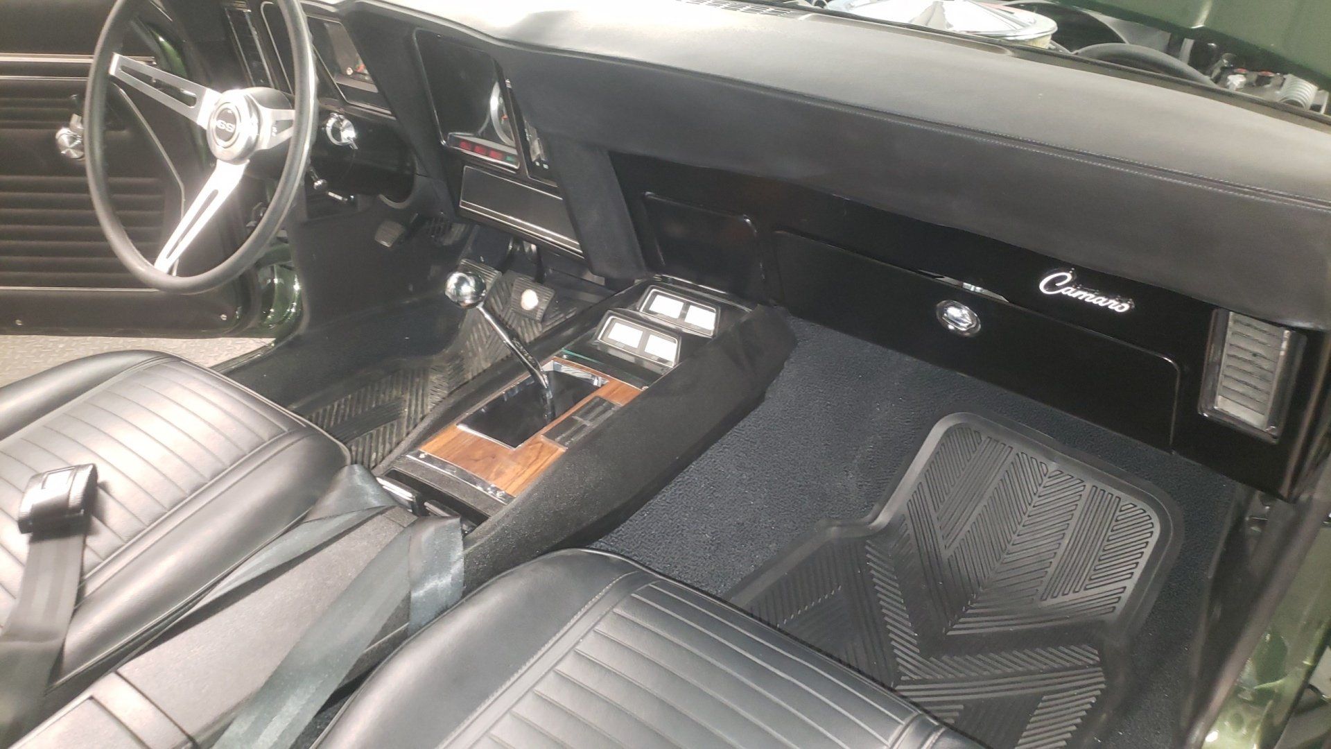 Interior view of a classic black Camaro with black seats, floor mats, and steering wheel, wood-grain console.