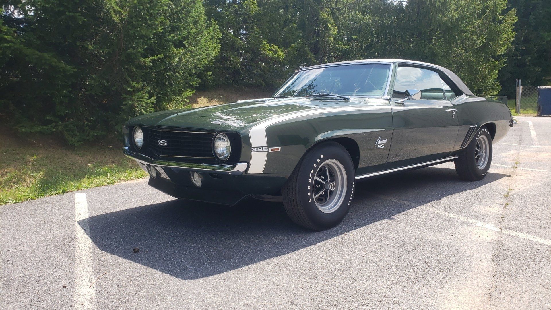 Green classic Camaro parked in an asphalt parking lot with white stripes and trees in the background.