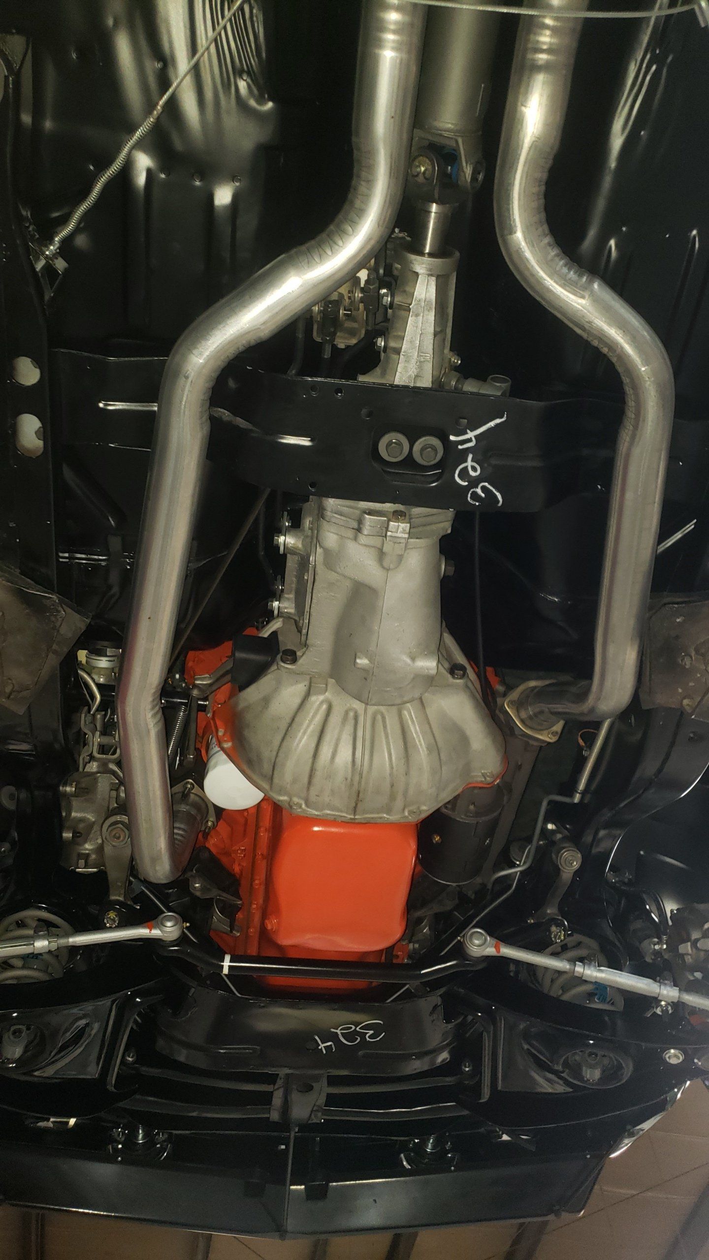 Undercarriage view of a classic car with orange engine, silver exhaust pipes, and a black painted frame.