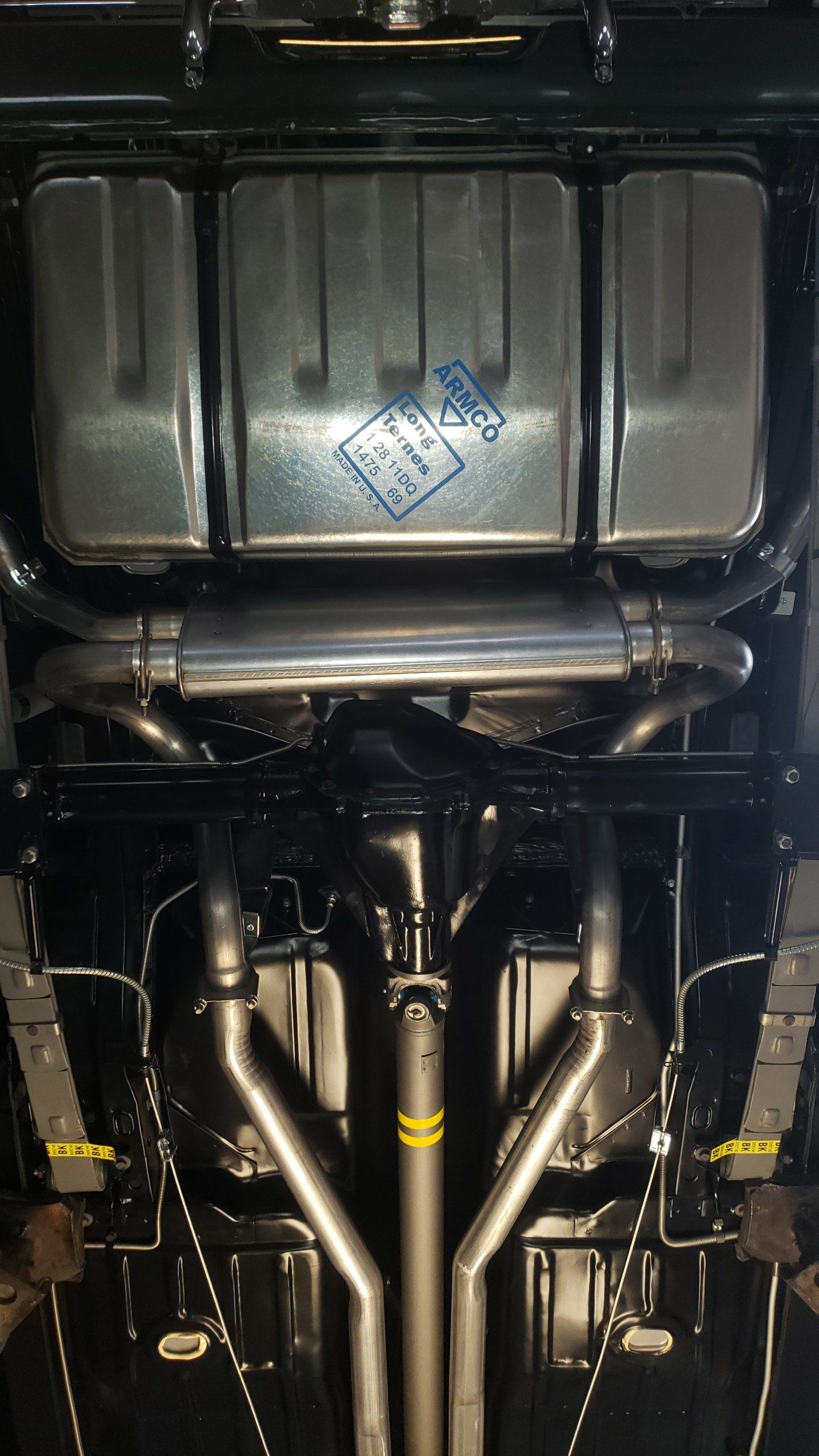 Underside of a classic car showing stainless steel exhaust system, fuel tank, and suspension components.