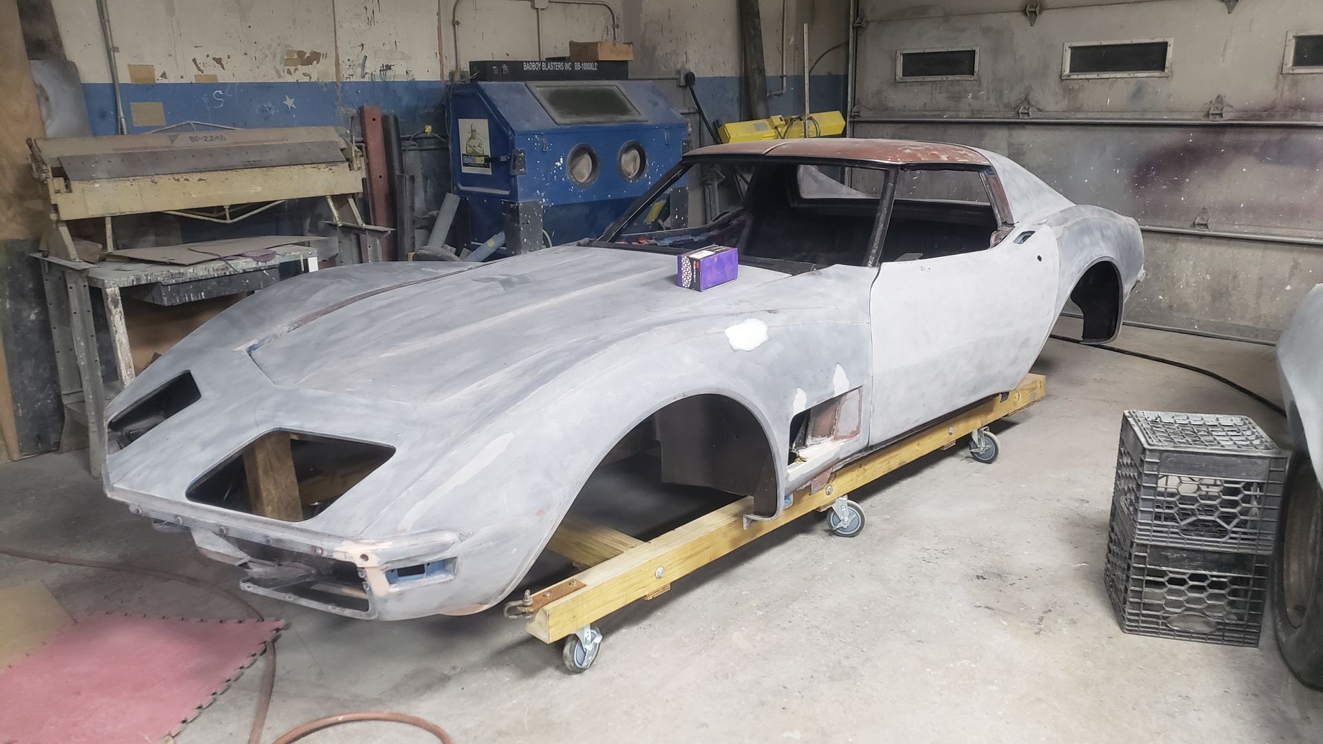 Classic Corvette car body on a wheeled platform in a workshop, undergoing restoration. Gray primer, exposed frame.