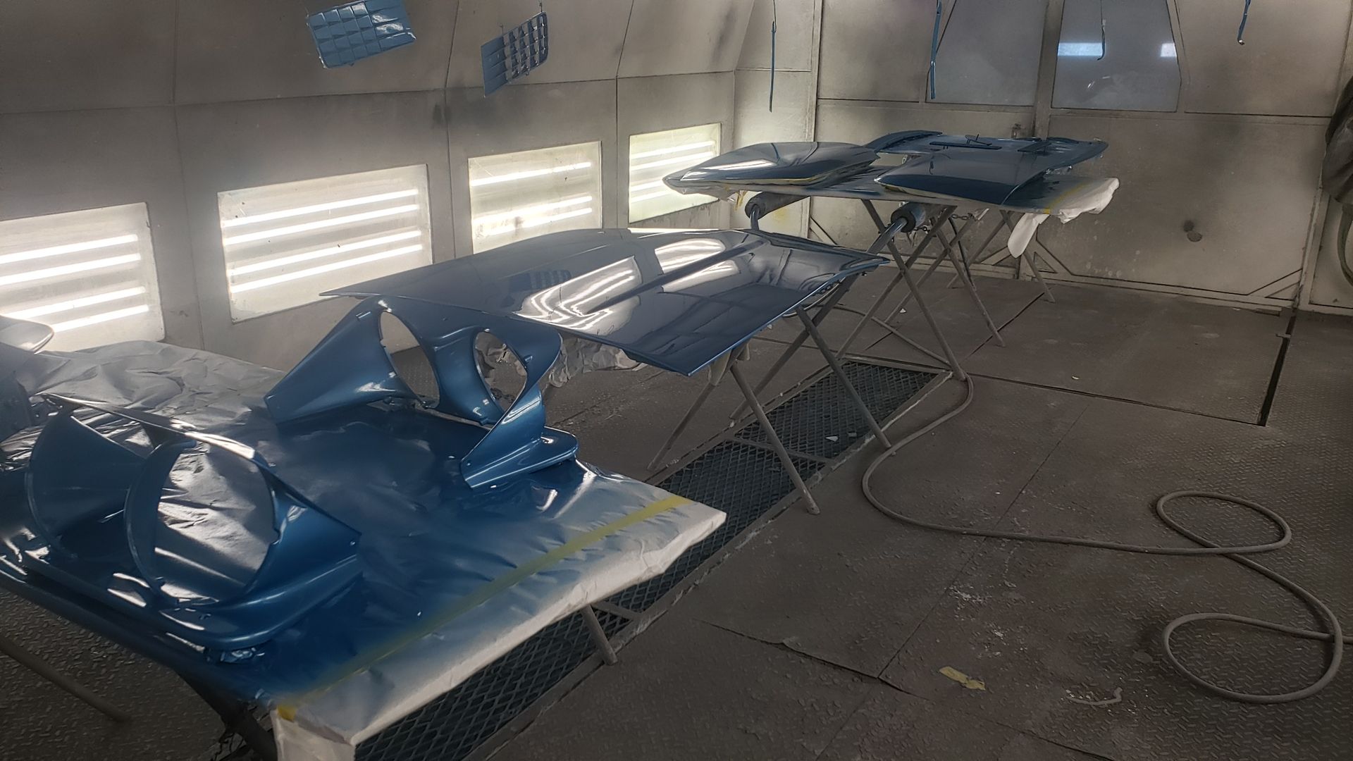 Blue car parts in a spray paint booth; parts on stands and workbench.