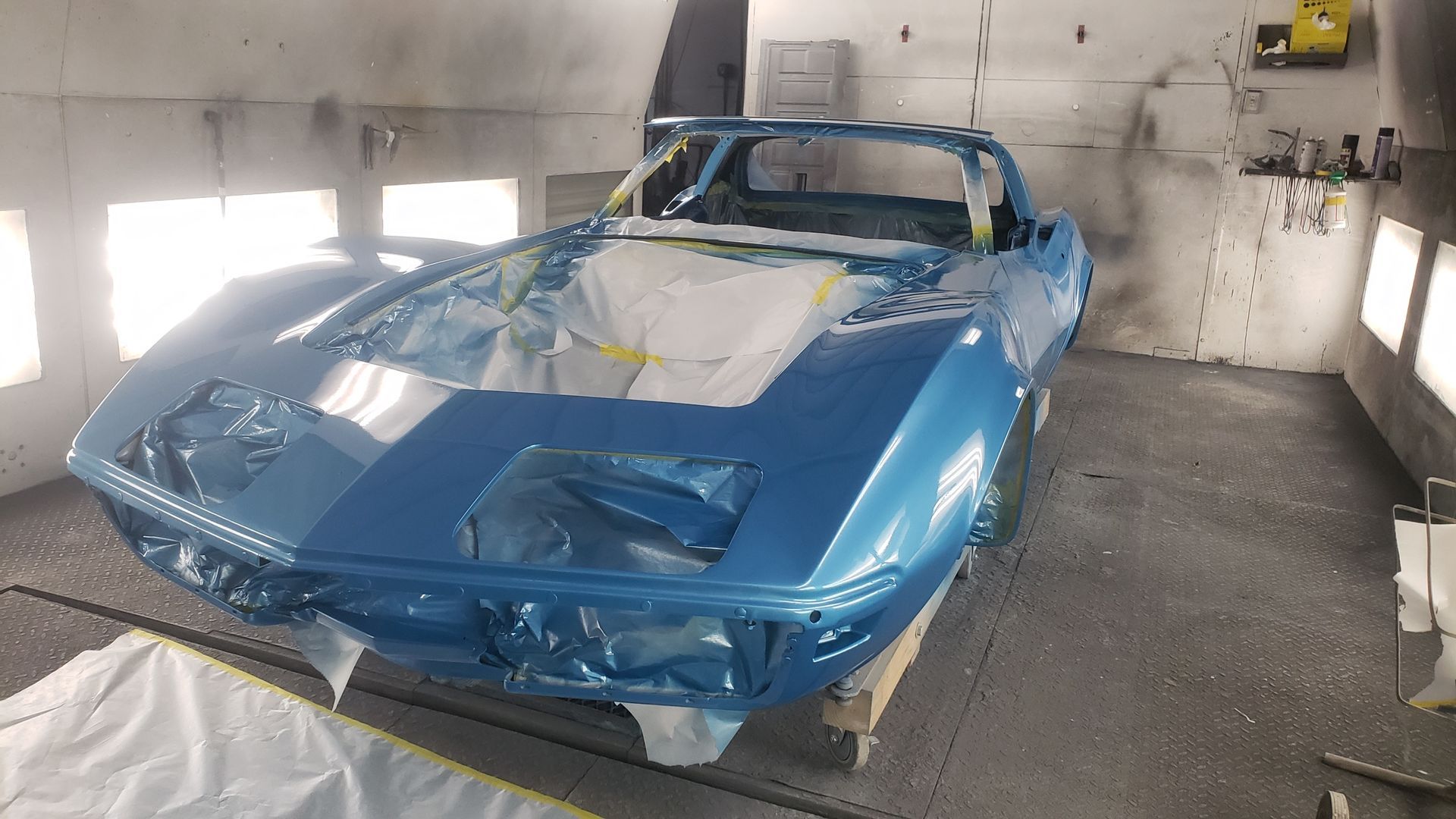 Blue classic Corvette in a paint booth, covered in plastic for protection.