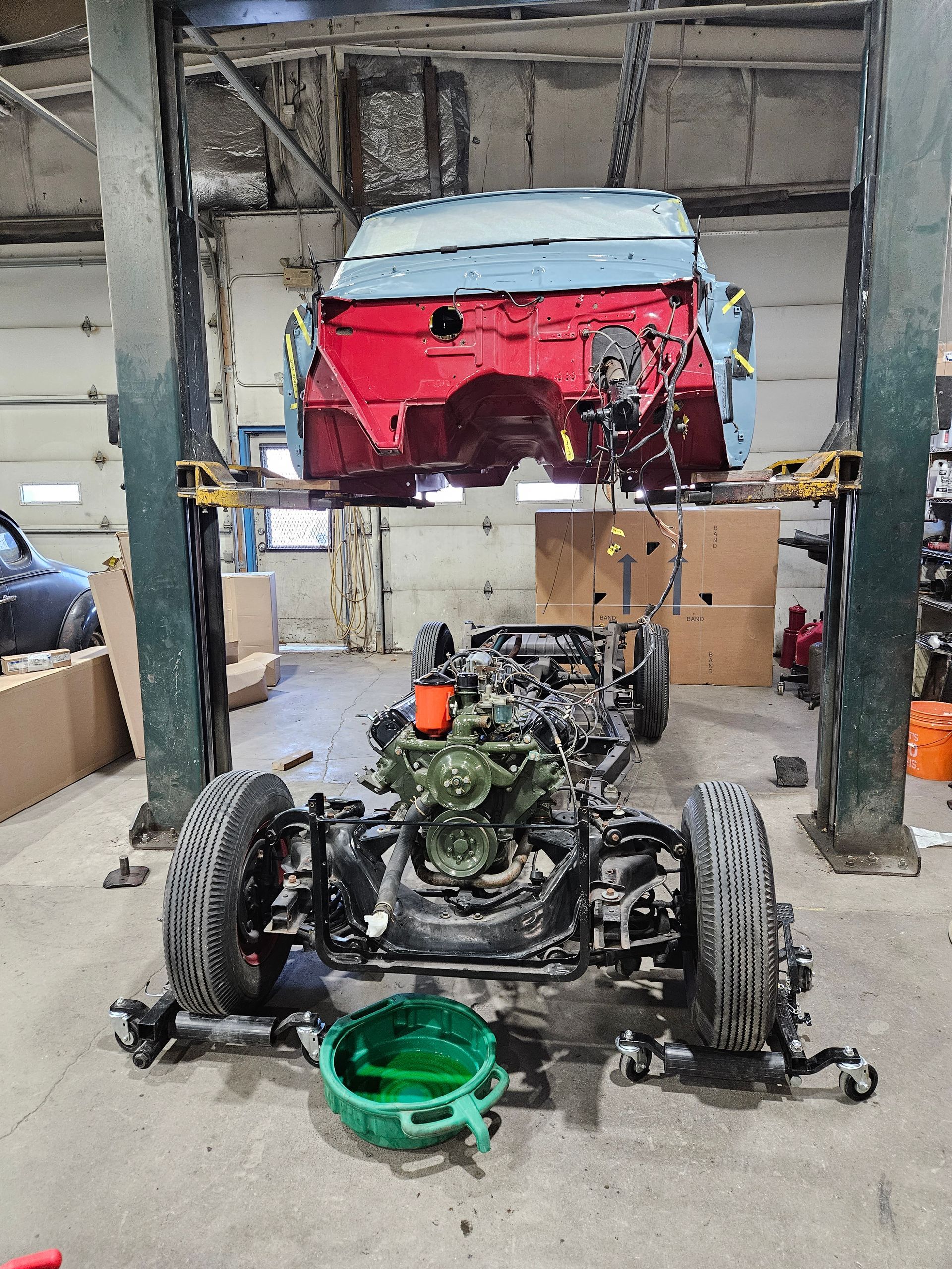 Car frame and body separated in a garage, body raised on a lift. Engine in the frame, green fluid in a pan.