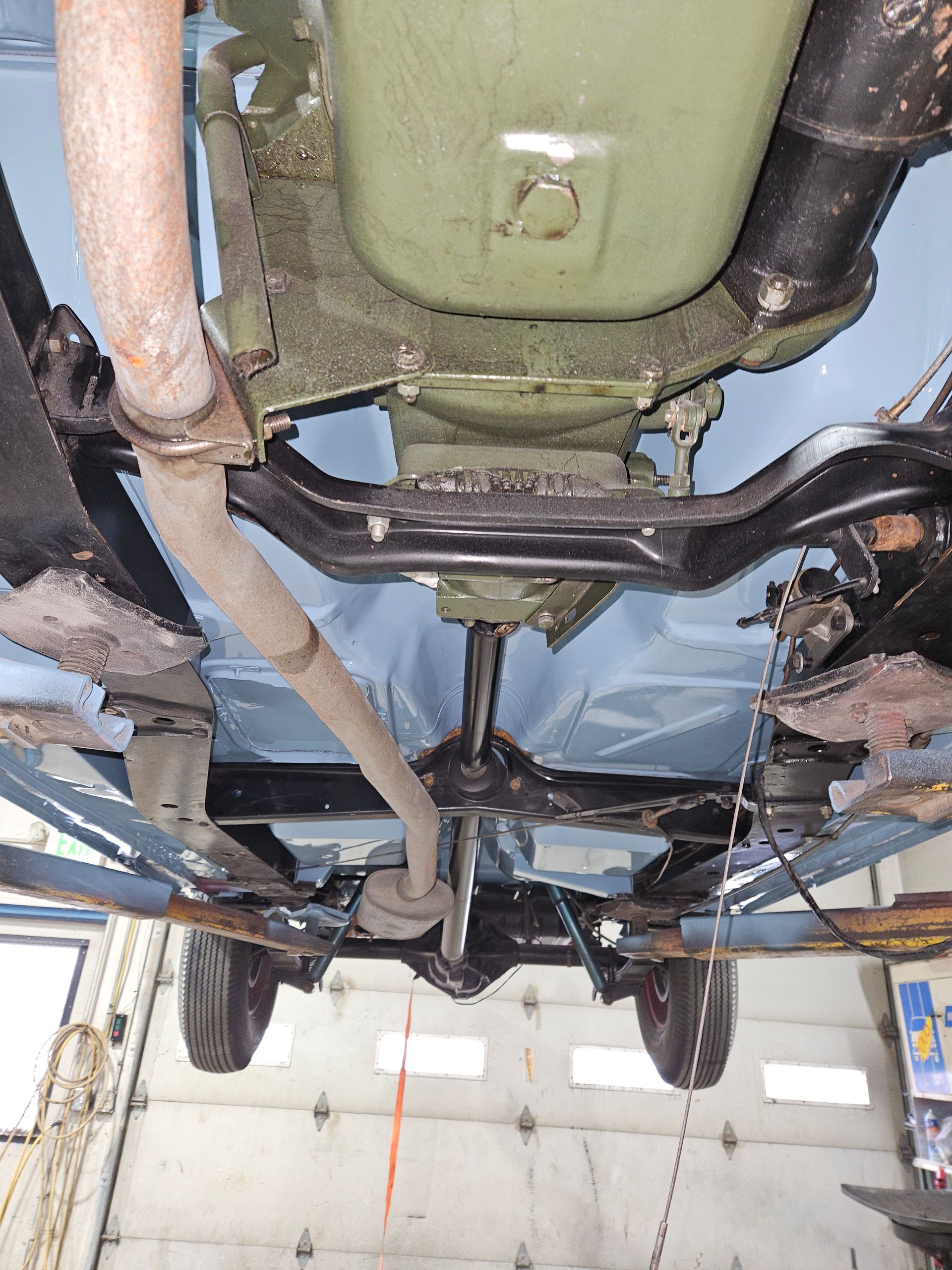 Underside of a light blue vintage car on a lift; visible are mechanical components, exhaust, and painted floorboards.