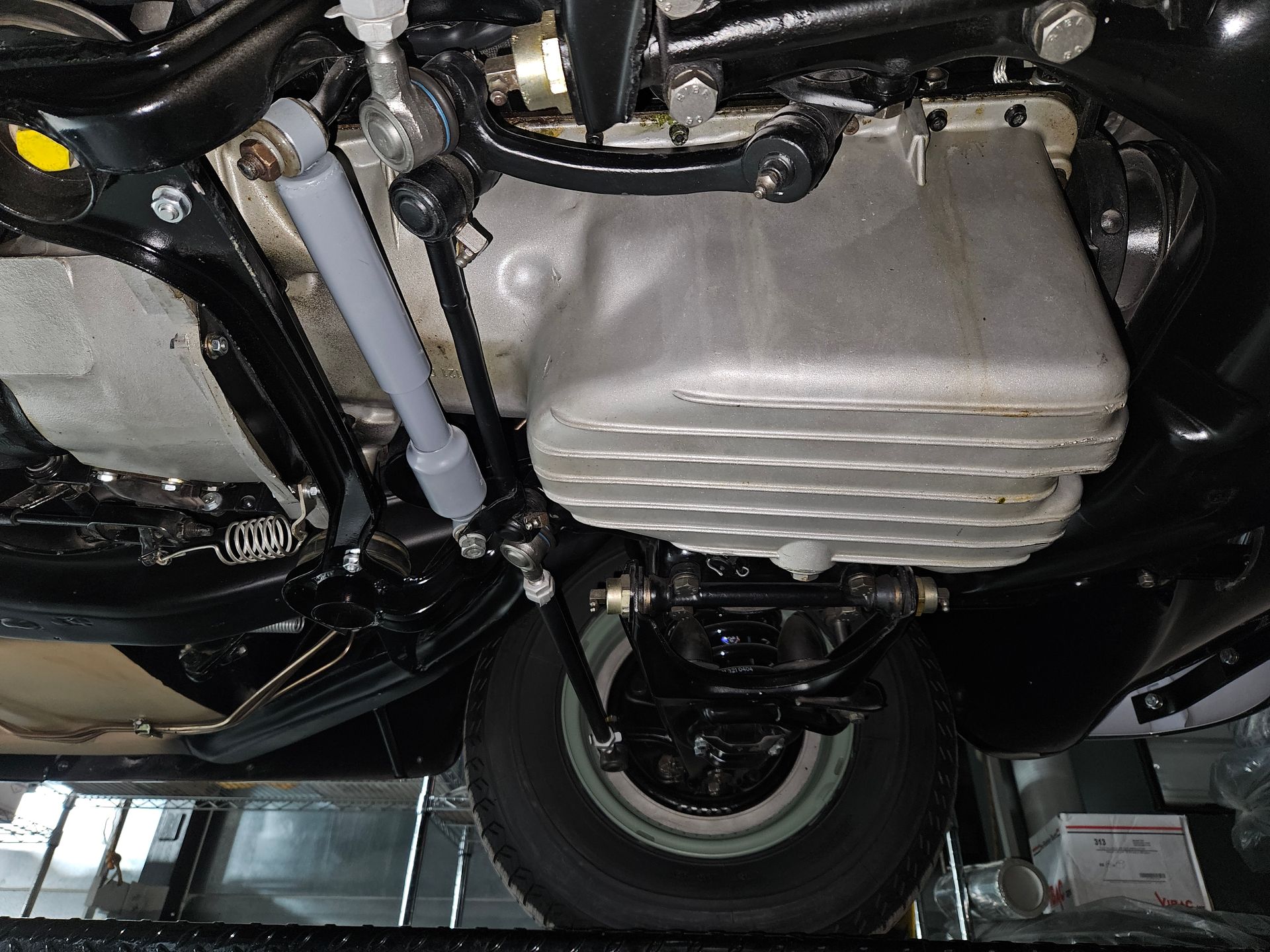 Underside view of a vehicle, showing engine oil pan, suspension components, and a wheel. Black and silver details.