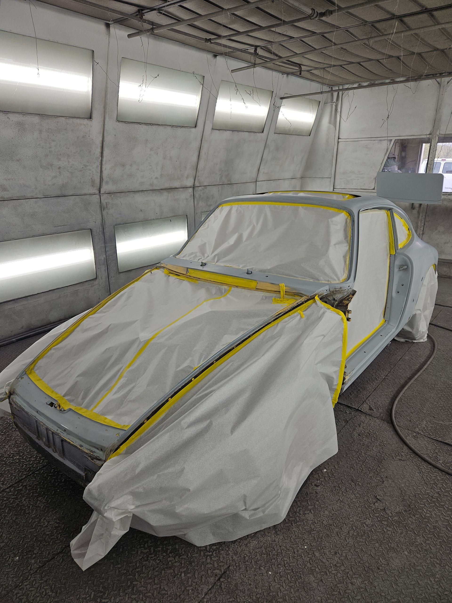 Car masked for painting inside a spray booth. Covered in white sheeting with yellow tape outlining areas to be painted.