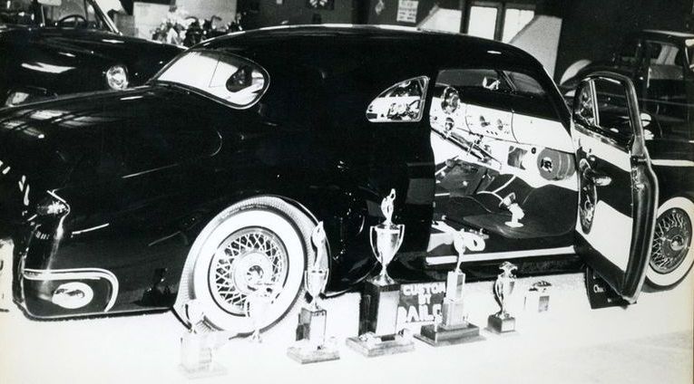 Black custom car with open doors, white walls, and trophies on display at a car show.