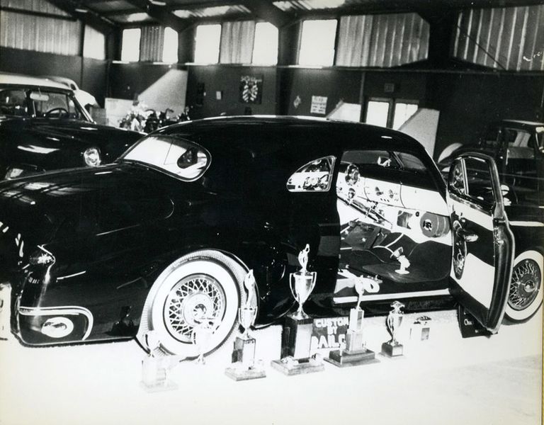 Black classic car with open door, displayed at a show with trophies and white wall tires.