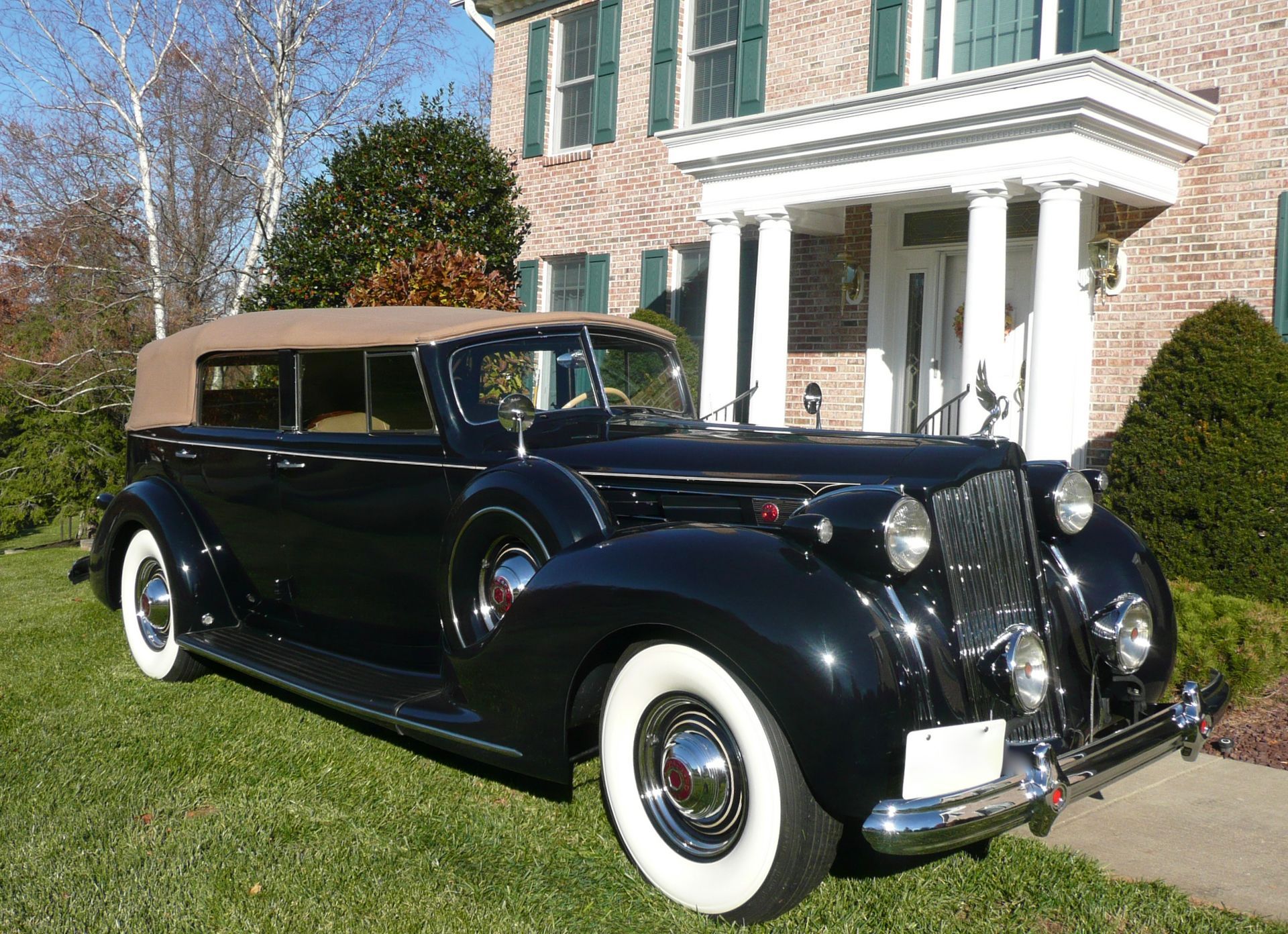 Black vintage Packard car with tan top parked in front of a brick house with white columns.