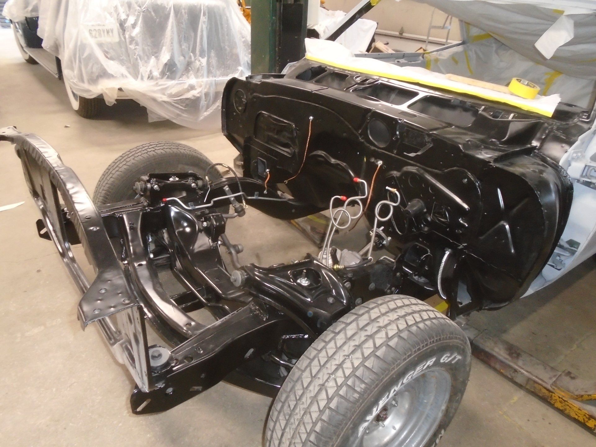 Black car chassis undergoing restoration with tires mounted. Shop setting.