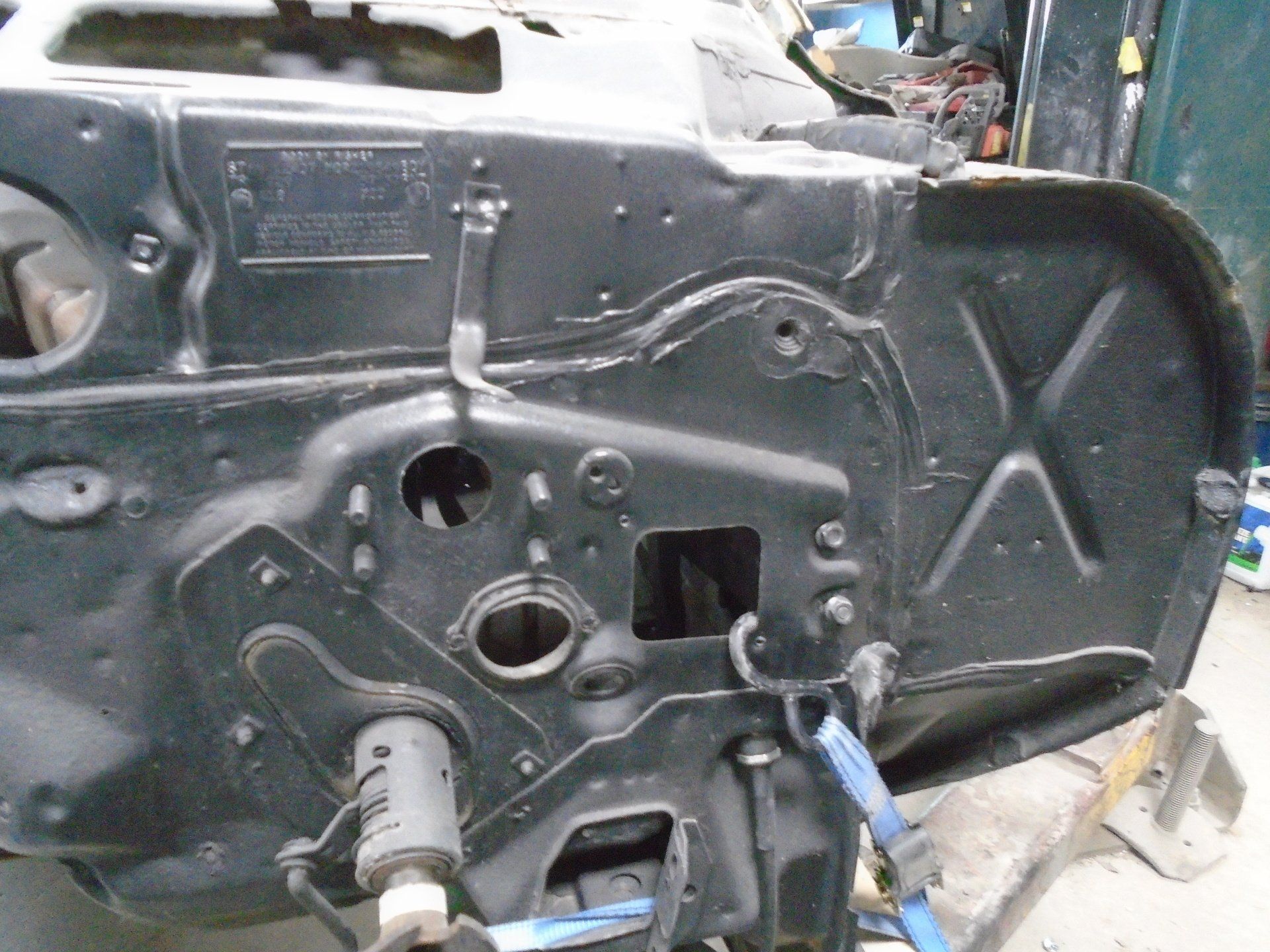 Black car engine compartment, various holes and brackets visible, blue strap.