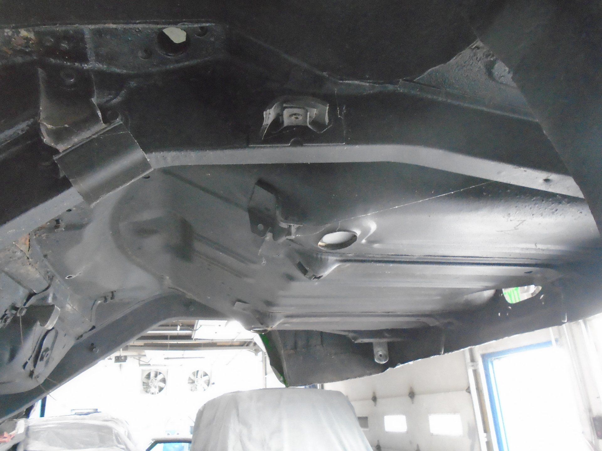 Underside of a black car chassis, showing structural components and floor pan, in a workshop setting.