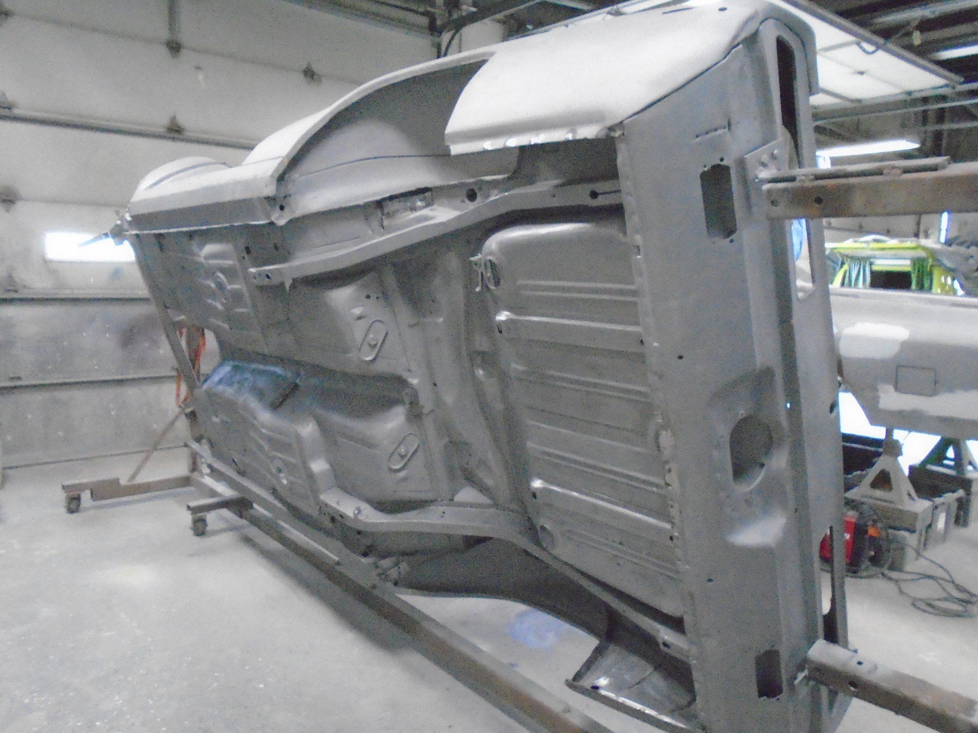 Underside of a car body shell, gray primed, mounted on a rotisserie in a shop.